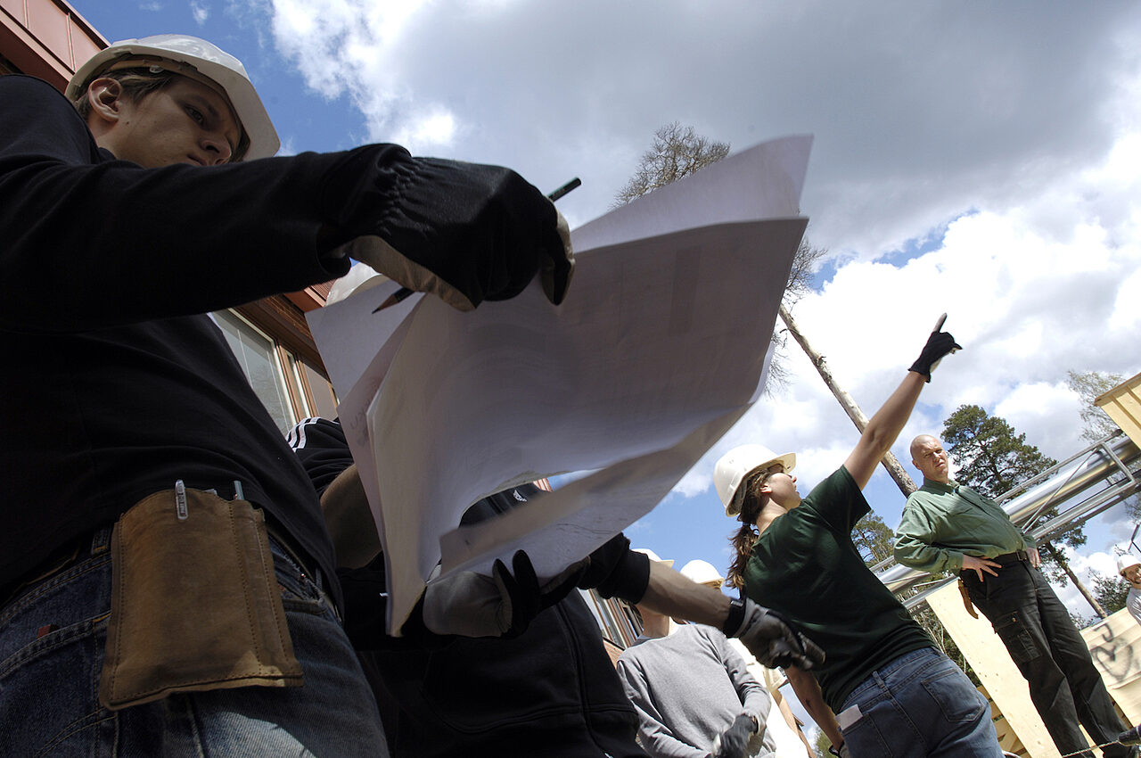 Näkymä alaviistosta ihmisiin, joista osa katsoo kädessään olevia papereita. Ihmisillä on päässään kypärät.