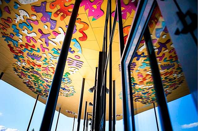 Talon katos, jota pitää ylhäällä mustat ohuet pylväät. Katoksen alapinta on lämpimän keltainen ja siinä on kirjavia ja abstrakteja kohokuvia.