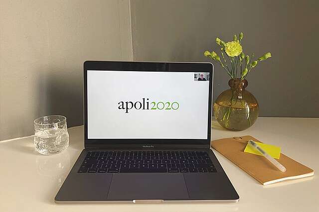 Pöydällä vesilasi, kannettava tietokone, jonka näytöllä lukee apoli2020, sekä kukkamaljakko ja muistiinpanovälineet.