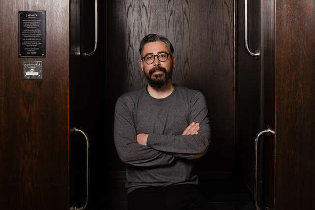 Kuvassa on mies, joka katsoo kameraan. Miehellä on harmaa paita.