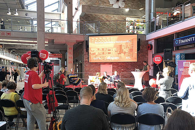 SuomiAreena-keskustelu lavalla ison näytön äärellä. Keskustelua seuraavat yleisö ja kuvaaja.