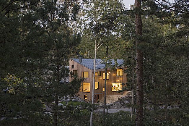 Puinen, teräväkulmainen ja geometrinen kolmikerroksinen talo näkyy havumetsän keskellä. Puisen talon suurista ikkunoista hohkaa lämmintä valoa.