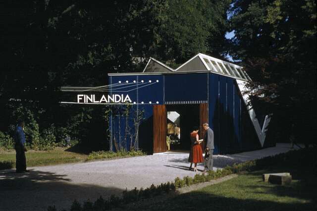 Sininen Finlandiapaviljonki luonnossa, siististi pukeutunut pari odottaa sen ulkona.