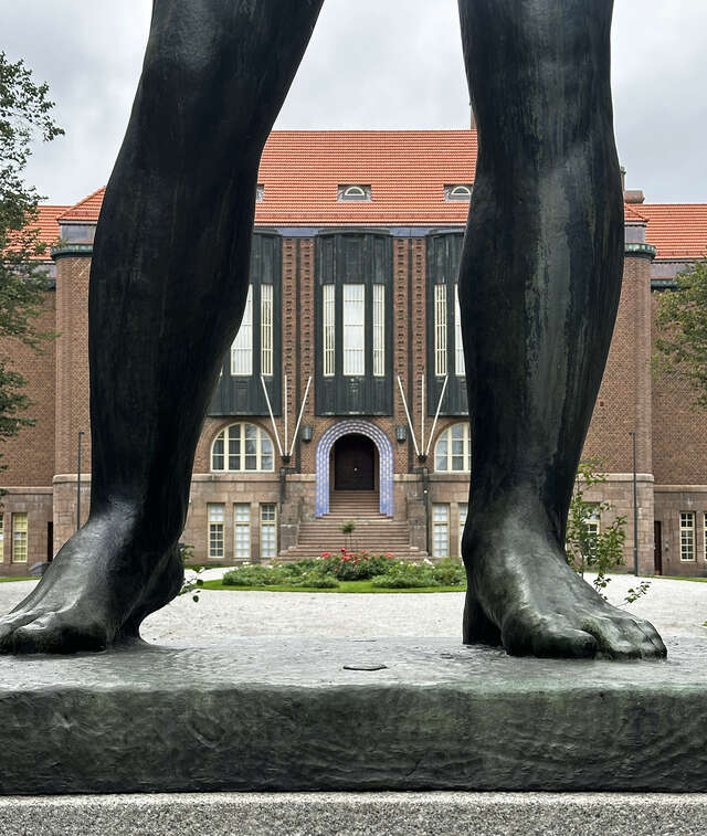 Etualalla olevan patsaan jalkojen välistä näkyy ruskeatiilinen komea talo