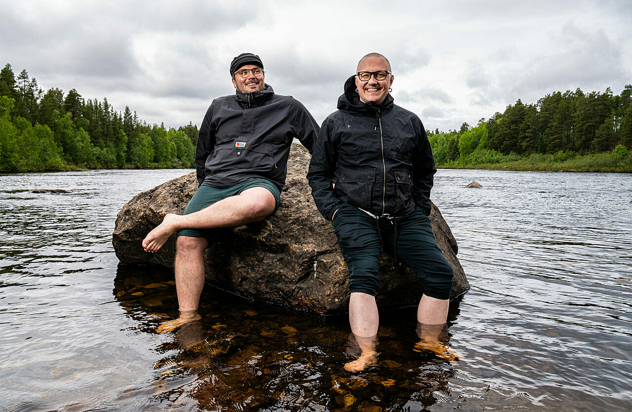 Two men sitting on a rock surrounded by water, with forest in the background.