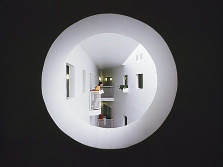 Pyöreästä aukosta näkyy pieni parveke, jolla seisoo ihminen. Parveke on talon sisällä ja sitä vastapäätä on seinä, jossa on ikkunoita.