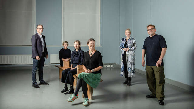 Two men and four women, some posing standing, some sitting in a lofty room.