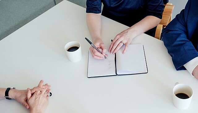 Kolme ihmistä kokoontuneena saman pöydän ääreen. Kaksi juo kahvia ja yksi kirjoittaa muistiinpanoja vihkoon.