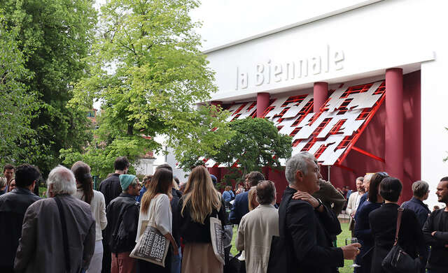 A crowd in front of a white building with text "la Biennale" and a red and metal installation serving as a canopy.