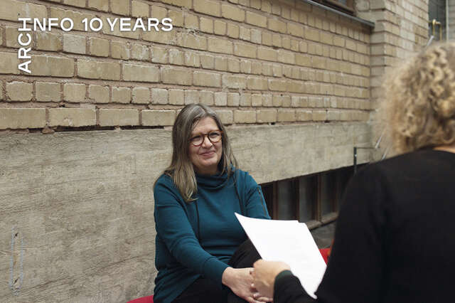 A woman in a turquoise sweater is sitting next to yellow-brick wall talking to a woman with curly blond hair.