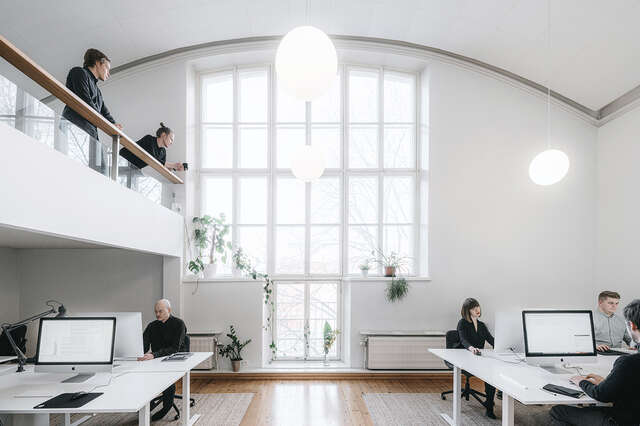 Sisäkuva korkeasta valkoisesta tilasta, taustalla kaari-ikkuna, vasemmalla parvi. Ihmisiä työskentelee työpöytien ja tietokoneiden ääressä.