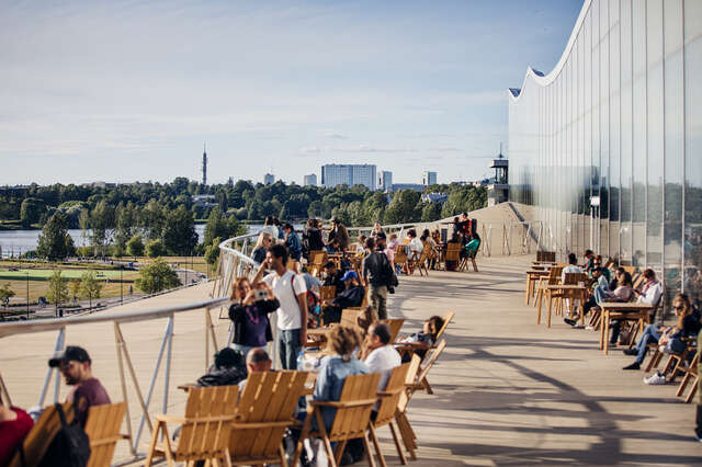 People on a summer roof terrace, on the right a glass wall with a corrugated roof.