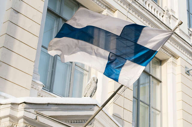 Suomen lippu liehuu vanhan rakennuksen seinään kiinnitetyssä lipputangossa.