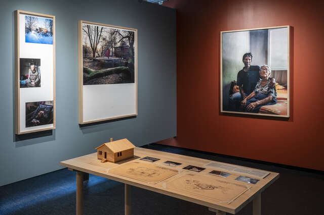 An exhibition showroom with several photagraphs hanging on walls and a table that has a scale model of a wooden building and drawings.