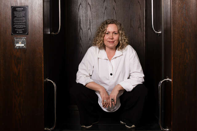 Nainen, joka katsoo suoraan kameraan. Hänellä on valkoinen paita.