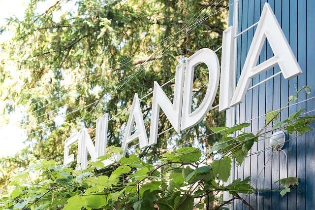 Letters forming FINLANDIA hanging from wires, surrounded by vines, on a backdrop of a blue wood facade.