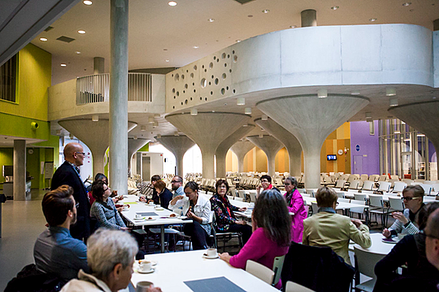 Näkymä salista, jossa ihmisiä istuu pöytien ääressä. Yksi henkilö seisoo ja moni pöydän ääressä istujoista katsoo häntä kohti.