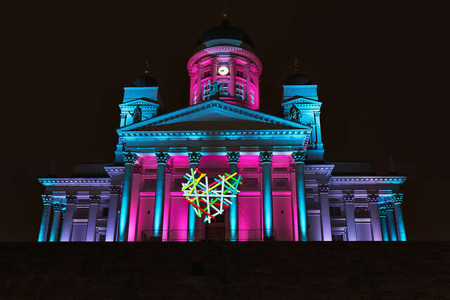 A large church lit with blue, lilac and pink lights. A heart is hung in front of the church's front doors.