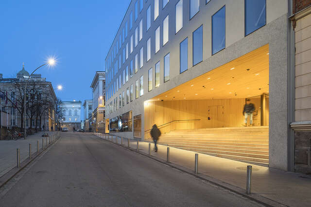 Iltakuva, nouseva katu, oikealla valoisa puupäällysteinen sisäänkäyntisyvennys valkoisessa talossa, jossa paljon ikkunoita.