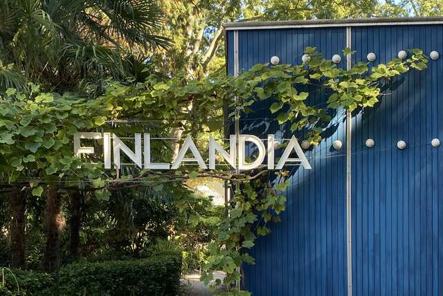 A sign that says Finlandia in large letters in front of a blue building and trees.