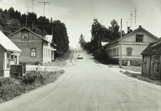 A black-and-white photo of wooden houses along a gravel road with a car descending the slope.