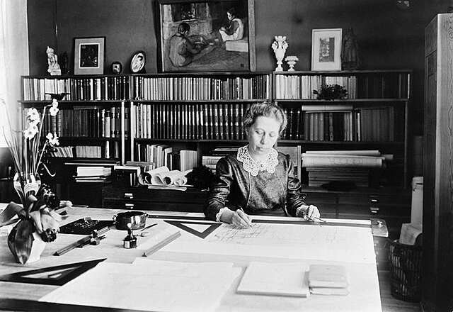 A woman sits on a table and looks at a large piece of white paper on the table.