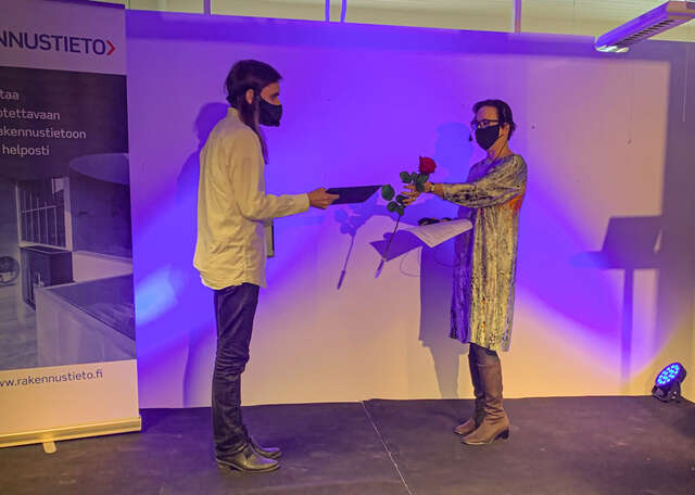 Lars-Erik Mattilalle ojennetaan kunniakirja ja ruusu lavalla. Palkinnonsaajalla ja palkinnon antajalla on molemmilla kasvomaskit.