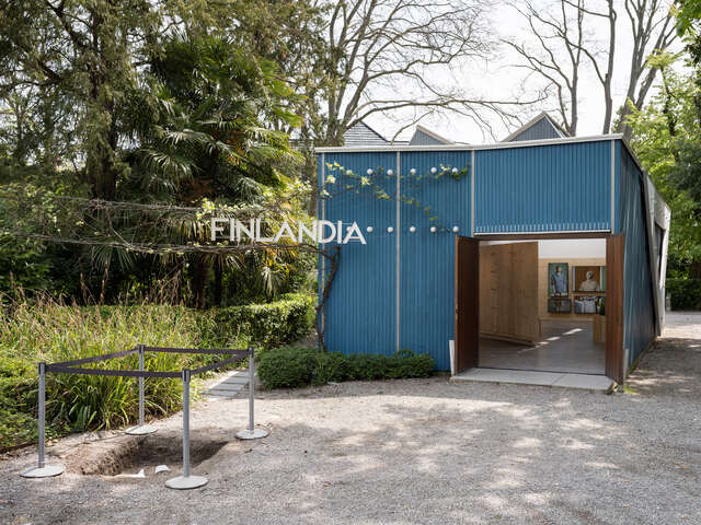 Blue wooden pavilion with doors open, inside an exhibition. In the front ground, a fenced-off excavation