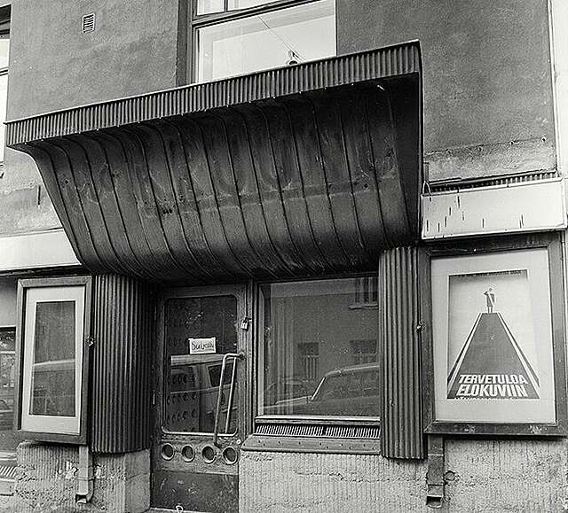 Picture of a façade with an arched canopy and an external door flanked by posters.