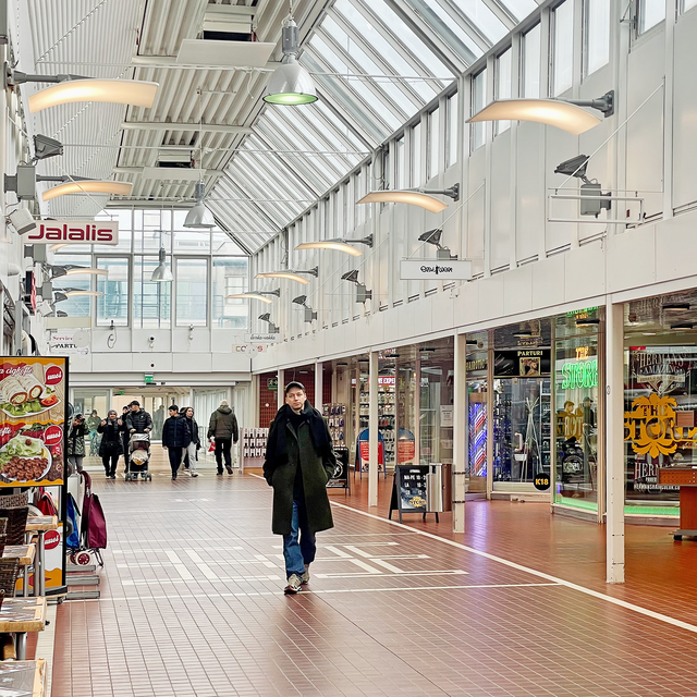 Mustatakkinen lippalakkipäinen mies kulkee kauppakeskukselta näyttävässä käytävässä