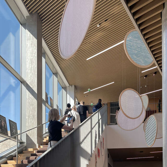 Ihmisiä nousemassa ylös portaita valoisassa, korkeassa tilassa, jonka katosta roikkuu pyöreiden levyjen installaatio.