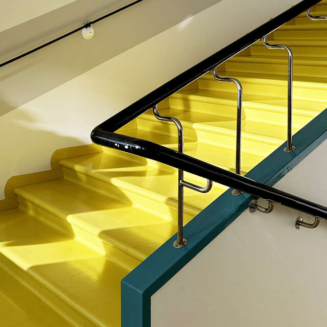 Yksityiskohta keltaisen portaikon mustasta kaiteesta, jossa on kulumisen jälkiä.