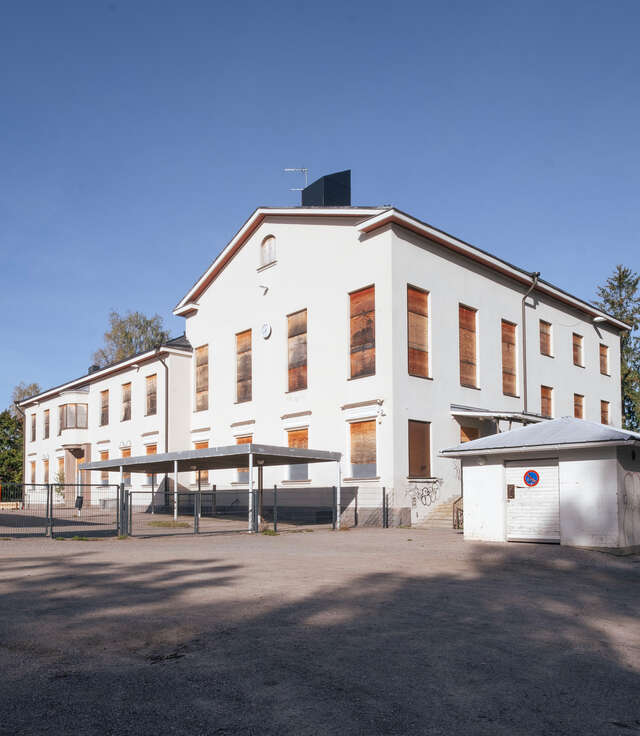 Vanha hylätty kansakoulu valkoisilla julkisivuilla, jonka ikkunat on peitetty vanerilla