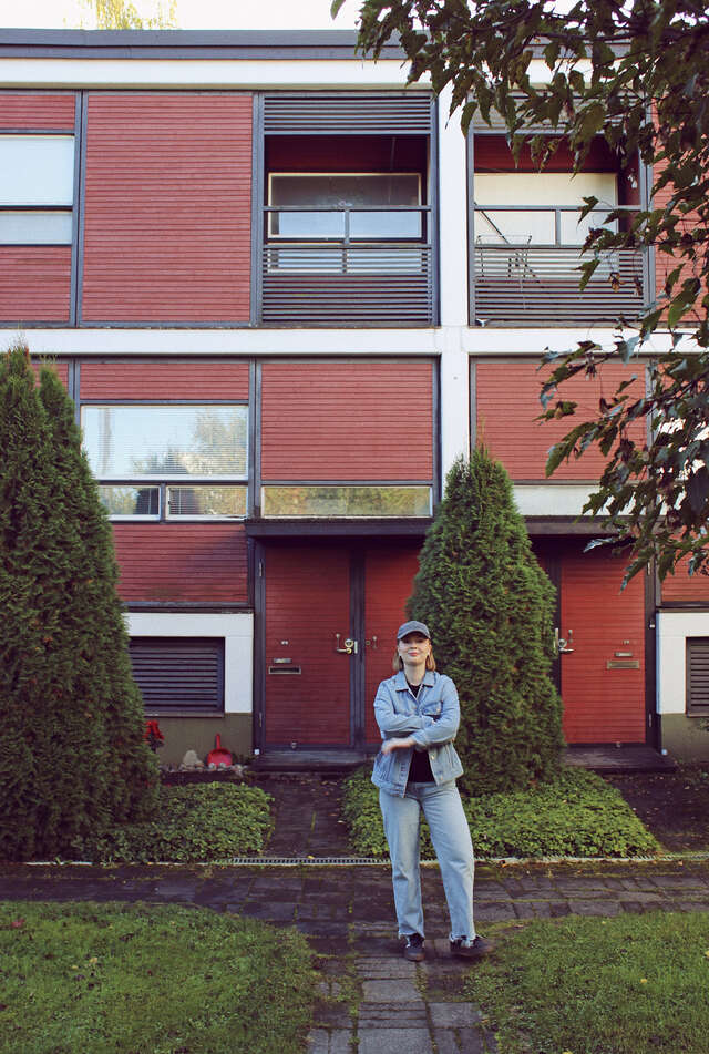 Nainen seisomassa punaisen rivitalon edessä. Pihassa näkyy pensaita.