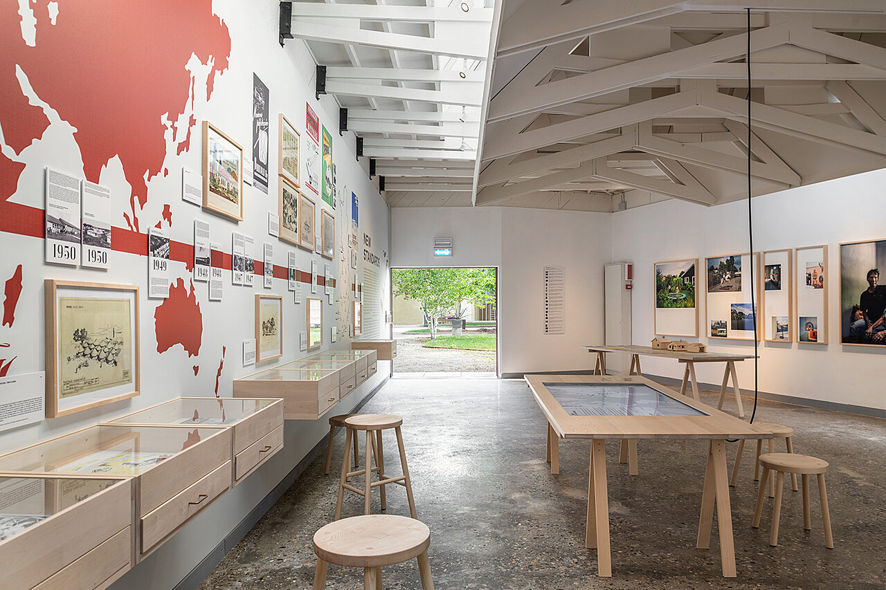 Sisäkuva paviljongista, vasemmalla seinällä punainen kartta ja viiva sekä kehystettyjä vanhoja kuvia. Keskellä pöytä, johon on upotettu näyttö. Oikealla seinällä koko seinän kokoinen valokuvateos, joka koostuu useasta valokuvasta ja kehyksestä.